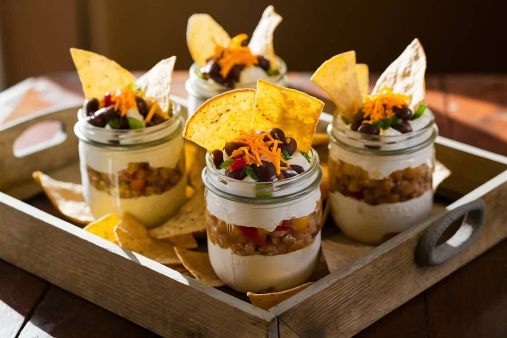 Individual servings of taco dip recipe in small mason jars, garnished with fresh vegetables and cheese, served with tortilla chips.